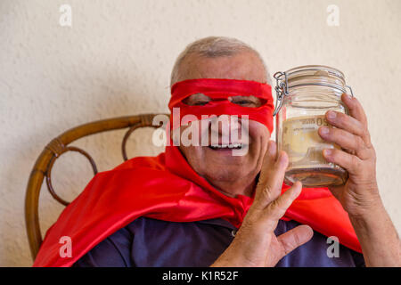 Vieil homme habillé en super héros, en souriant, assis sur chaise en osier et tenant un pot plein de billets sur fond de mur jaune Banque D'Images