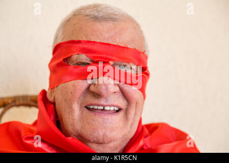 Vieil homme habillé en super héros smiling while sitting on Wicker Chair et tenant une rose rouge sur fond de mur jaune Banque D'Images