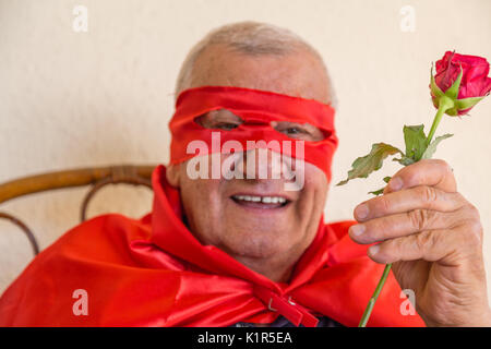 Vieil homme habillé en super héros smiling while sitting on Wicker Chair et tenant une rose rouge sur fond de mur jaune Banque D'Images