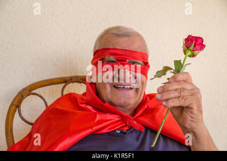 Vieil homme habillé en super héros smiling while sitting on Wicker Chair et tenant une rose rouge sur fond de mur jaune Banque D'Images