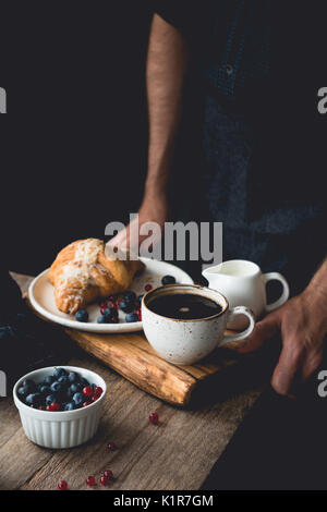 Petit déjeuner sur un plateau : croissants, café, crème et petits fruits. man hands holding plateau en bois avec petit déjeuner Banque D'Images