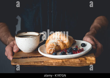 Petit déjeuner sur un plateau : croissants, café et petits fruits. man hands holding plateau en bois avec petit déjeuner Banque D'Images