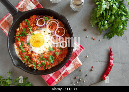 Shakshuka dans une poêle en fonte. Style moyen-orientale oeufs pochés dans une sauce tomate et du poivre avec le persil, l'oignon et les épices sur fond de béton gris. Banque D'Images