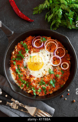 Shakshuka dans une poêle en fonte. Style moyen-orientale oeufs pochés dans une sauce tomate et du poivre avec le persil, l'oignon et les épices sur fond de béton gris. Banque D'Images