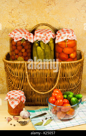 Des conserves de légumes (tomates, concombres, tomates cerises) dans un bocal en verre sur panier en osier, des légumes frais (tomates, concombres) dans bol en verre, G Banque D'Images