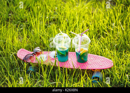 Deux cocktails de fruits debout sur la planche à roulettes. Les lunettes de soleil se coucher sur la planche à roulettes. Skate se trouve dans l'herbe verte. Une boisson rafraîchissante. Banque D'Images