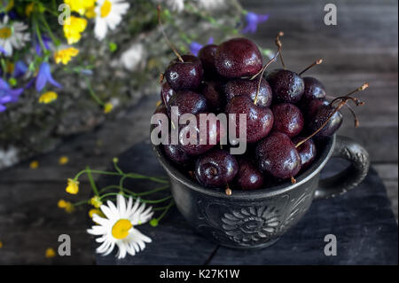 Cerises juteuses mûres dans un pot en argile avec les marguerites et les cloches dans un style rustique. Banque D'Images