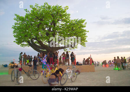 Brûleurs se réunir autour d'un arbre géant de la sculpture au coucher du soleil sur la playa comme le désert Burning Man Festival annuel ferme le premier jour de la semaine 27 août 2017 Black Rock City, Nevada. Le festival annuel attire 70 000 visiteurs dans l'une des régions les plus isolées et les déserts inhospitaliers en Amérique. Banque D'Images