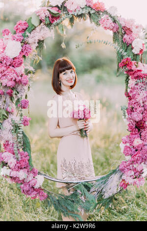 Le portrait en gros plan de la demoiselle souriante avec les fleurs debout derrière le mariage de pivoines arch dans le domaine ensoleillé. Banque D'Images