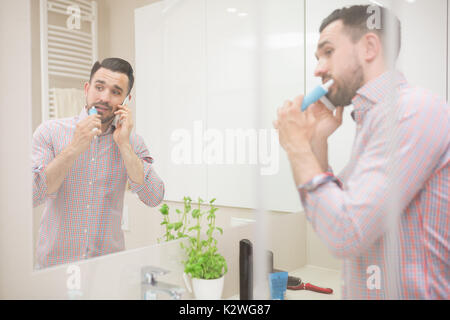 Homme debout dans la salle de bain, se brosser les dents et d'avoir l'appel téléphonique. Il est à l'aide de brosse à dents électrique, à l'écart et portant chemise décontractée avec retroussé Banque D'Images