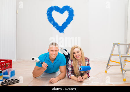 Portrait of Young Couple with Paint Roller Lying On Floor In New Home Banque D'Images