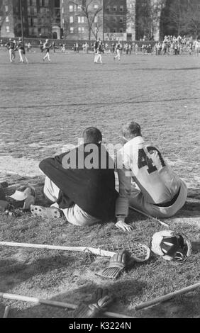 Sépia photographie candide de deux co-capitaines de l'équipe de crosse à l'Université Johns Hopkins, Joe Sollers et Byron Forbush, assis à partir de la ligne de touche sur l'herbe avec dos tourné, regarder la fin du match, avec des gants et des bâtons de crosse éparpillés derrière eux, Baltimore, Maryland, juin, 1951. Banque D'Images