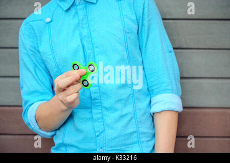 Yong élégant garçon sur l'azur shirt et jeans bleu hand holding gadget populaire fidget spinner. L'homme jouant avec spinner vert à l'extérieur sur la brique Banque D'Images