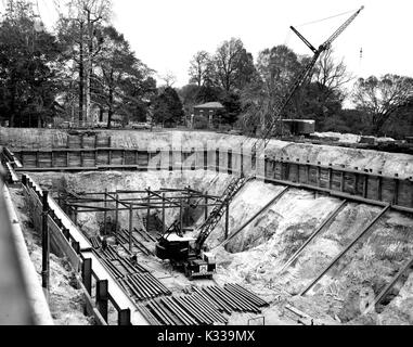 Au cours des premières étapes de la construction de la Milton S Eisenhower Library à l'Université Johns Hopkins, une grande grue s'étend de la fondation du bâtiment, où des poutres en acier sont empilés et disposés de façon à créer une structure au-dessus du sol, avec des bâtiments du campus et les arbres bordant les motifs du projet, et des matériaux de construction et les véhicules stationnés tout environ, Baltimore, Maryland, 1963. Banque D'Images