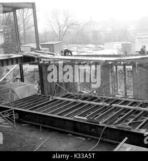 Au cours des premières étapes de la construction de la Milton S Eisenhower Library à l'Université Johns Hopkins, de grandes poutres en acier sont empilés et disposés de façon à créer la structure de Q et M niveaux, avec les travailleurs debout sur des échafaudages et de grandes planches en bois, plusieurs voitures et camions stationnés sur le terrain herbeux en face, et la rue Charles visibles à l'horizon à travers le brouillard, Baltimore, Maryland, 1963. Banque D'Images