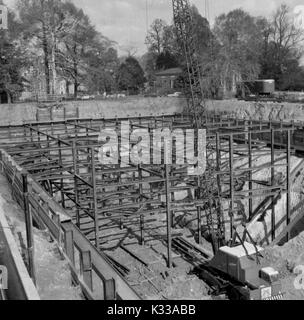 Au cours des premières étapes de la construction de la Milton S Eisenhower Library à l'Université Johns Hopkins, poutres d'acier sont empilés et disposés de façon à créer le début d'une structure au-dessus du sol, de la fondation à l'intérieur d'une fosse de terre, à l'aide d'une grue et d'autres véhicules dans la distance, et d'arbres et de bâtiments du campus bordant le terrain du projet, Baltimore, Maryland, 1963. Banque D'Images