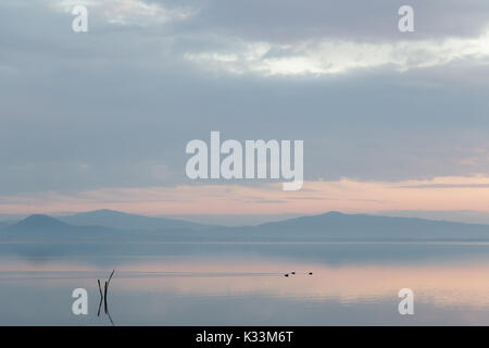 Un lac au crépuscule, avec une lumière douce, des couleurs chaudes, des collines et montagnes, certains canards sur l'eau et des poteaux en bois au premier plan Banque D'Images