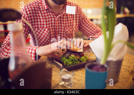 Une photo de young man eating big hamburger plein de légumes à de belles bar café. Banque D'Images