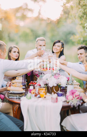 La photo en gros plan de les mariés et invités trinquer leurs lunettes et assis à la table pleine de nourriture délicieuse et de pivoines. Banque D'Images