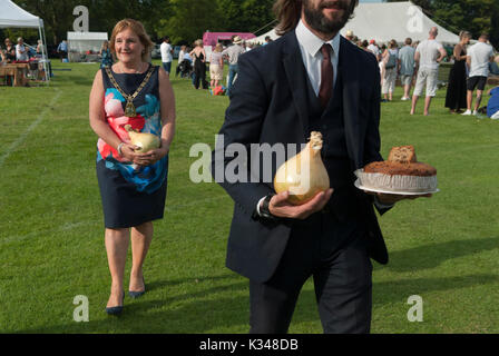 Maire de Bromley, Cllr Kathy Bance MBE avec des achats qu'elle a faits à la vente aux enchères de produits locaux et fabriqués localement au village de Cudham annuel Bank Holiday lundi show d'été et fête. Sa sécurité et son chauffeur avec un oignon surdimensionné et un gâteau fait maison mènent le chemin du retour à sa voiture officielle. Cudham, Kent, Angleterre 28 août 2017 2010s Royaume-Uni HOMER SYKES Banque D'Images