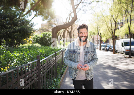 Une photo de jeune homme debout sur le trottoir avec un téléphone mobile dans ses mains. Il est souriant, heureux. Banque D'Images