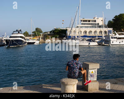 Un garçon local regarde les bateaux dans le port de Porto Christo. Le grand yacht de Rafa Nadal se trouve sur la gauche de la photo et le Club Nautico est au milieu. Le yacht de Rafa est le plus grand dans le port et a la position d'amarrage de premier choix comme il est un héros local. Banque D'Images