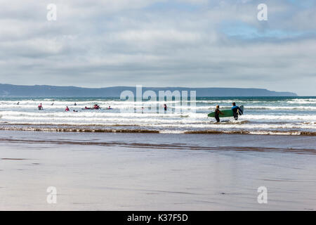 Deux surfers carrying leurs planches de surf dans la mer à Westward Ho !, Devon, Angleterre, Royaume-Uni, d'autres surfeurs à l'arrière-plan Banque D'Images