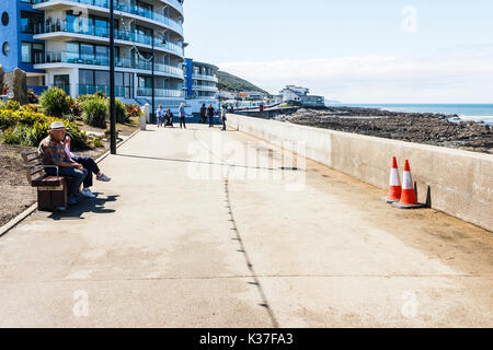 Un homme âgé dans hat assis sur un banc sur la promenade à Westward Ho ! Face à deux cônes de circulation par le mur de la mer, appartements de vacances dans l'arrière-plan Banque D'Images