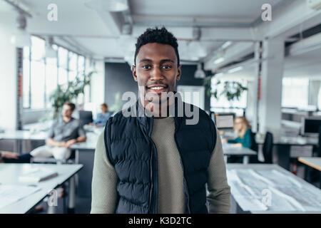 Portrait of happy young woman standing in office avec des gens assis en arrière-plan. African Male designer debout dans un bureau moderne. Banque D'Images