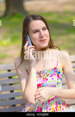 Portrait de jeune belle femme aux cheveux longs en été, parc girl wearing flower robe courte est à l'aide d'un smartphone tout en étant assis sur un banc Banque D'Images