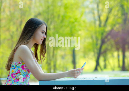 Portrait de jeune belle femme aux cheveux longs en été, parc girl wearing flower robe courte est à l'aide d'un smartphone tout en étant assis sur un banc Banque D'Images