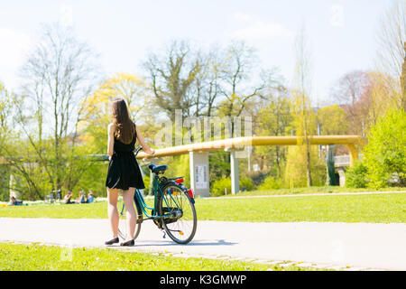 Vue arrière de la fille sur location portant sur noir robe courte. Jeune femme équitation le long de la route sur vert printemps parc extérieur. Jeune fille sportive équitation un bic Banque D'Images