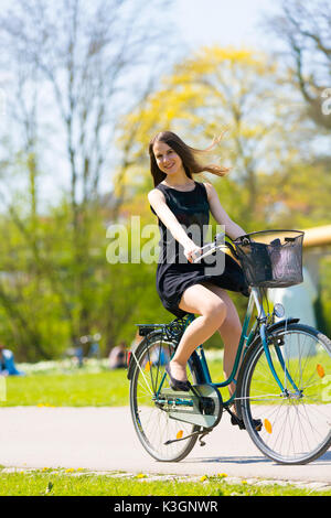 Vue Portrait of Girl sur location portant sur noir robe courte. Jeune femme heureuse équitation le long de la route sur vert printemps parc extérieur. Jeune fille sportive ri Banque D'Images