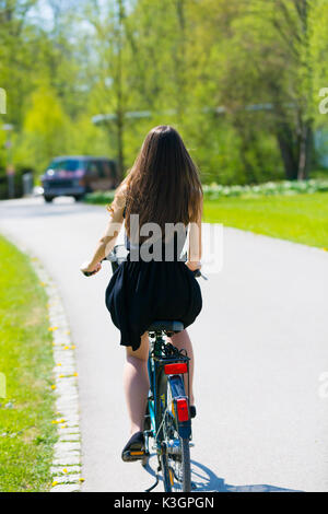 Vue arrière de la fille sur location portant sur noir robe courte. Jeune femme équitation le long de la route sur vert printemps parc extérieur. Jeune fille sportive équitation un bic Banque D'Images