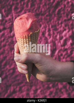 Main d'un garçon tenant un cornet de crème glacée aux fraises sur fond rose Banque D'Images