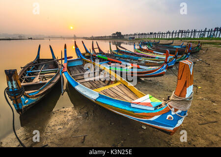 Amarapura, région de Mandalay, Myanmar. Bateaux colorés amarrés sur les rives de la lac Taungthaman au lever du soleil, avec l'U Bein bridge en arrière-plan. Banque D'Images