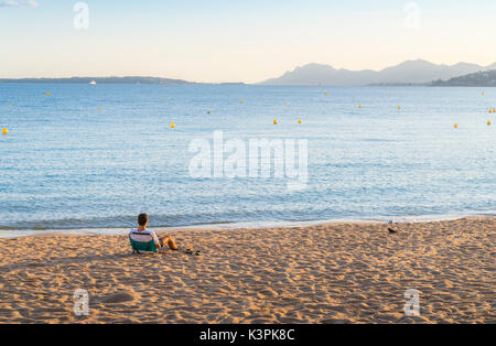 Un jeune adulte se repose sur une chaise longue de plage vide donnant sur une baie de la Côte d'Azur, France at sunset Banque D'Images