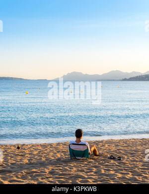 Un jeune adulte se repose sur une chaise longue de plage vide donnant sur une baie de la Côte d'Azur, France at sunset Banque D'Images