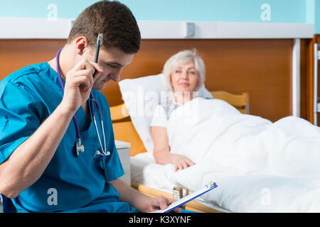 Médecin beau en uniforme avec phonendoscope sur son cou de mettre par écrit les plaintes de patient, qui est couché dans le lit d'hôpital à l'hôpital Banque D'Images