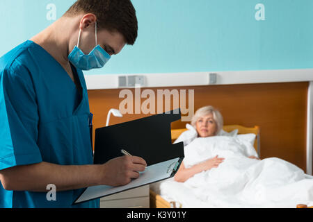 Beau jeune médecin en uniforme portant un masque de faire quelques notes dans le rapport au patient malade lors d'une visite de presse-papiers dans le quartier. Concept de soins de santé Banque D'Images