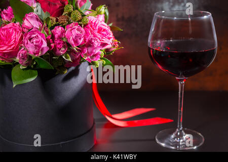 Bouquet de roses rose et rouge et ruban rouge dans une boîte noire circulaire près d'un verre à vin. Saint-Valentin et concept anniversaire Banque D'Images