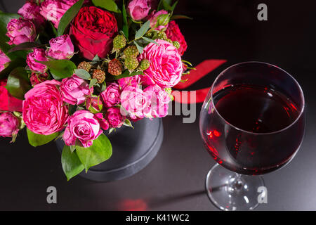 Bouquet élégant de rose et roses rouges et ruban rouge dans une boîte noire circulaire près d'un verre à vin. Saint-Valentin et concept anniversaire Banque D'Images