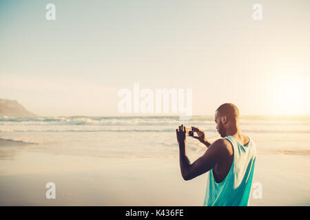 African man photographing on the beach en utilisant un téléphone mobile. Jeune mâle qui fait la photographie mobile à bord de la mer. Banque D'Images