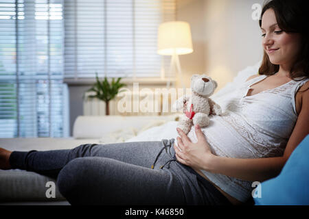 Happy pregnant woman sitting on couch et teddy bear. Banque D'Images