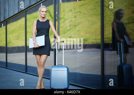 Face portrait of mature confident businesswoman holding laptop et poignée de valise. Banque D'Images