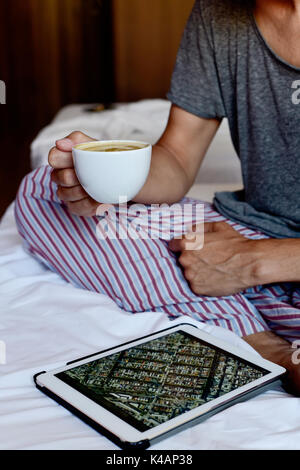Libre d'un jeune homme de race blanche en pyjama assis au lit après avoir un café tout est vérifier une adresse dans une carte dans sa tablette Banque D'Images