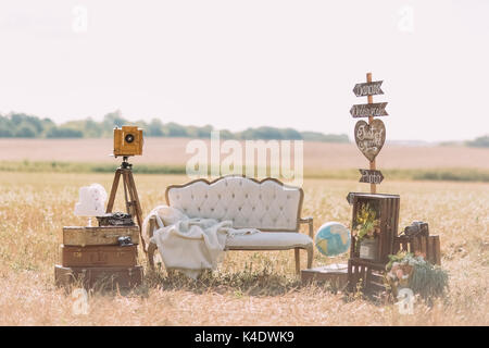 La composition de l'ancien canapé avec des étoffes à carreaux, plaques en bois avec les signes ''Wedding, dancing'', valises vintage avec globe et des fleurs dans le domaine ensoleillé. Banque D'Images