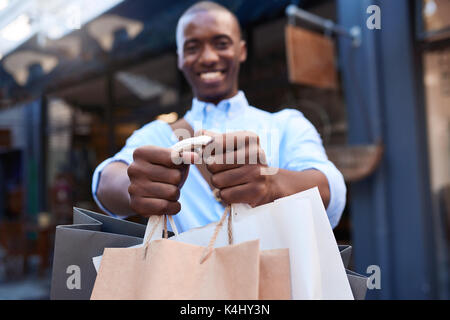 Portrait d'une jeune élégante african man smiling and holding up shopping bags tandis que les magasins dans la ville Banque D'Images