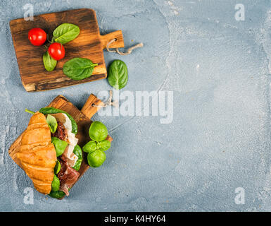 Croissant sandwich avec de la viande fumée jambon de Parme, tomates séchées, épinards frais et le basilic sur les petites planches de bois rustique en pierre sur fond gris texturé Banque D'Images