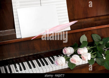 Composition de roses, rose pâle et blanc papier musical feuille blanche avec plume rose sur brown piano. Banque D'Images